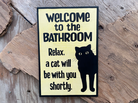 Sign with text 'Welcome to the Bathroom, Relax, a cat will be with you shortly.' and a black cat illustration on a wooden background.